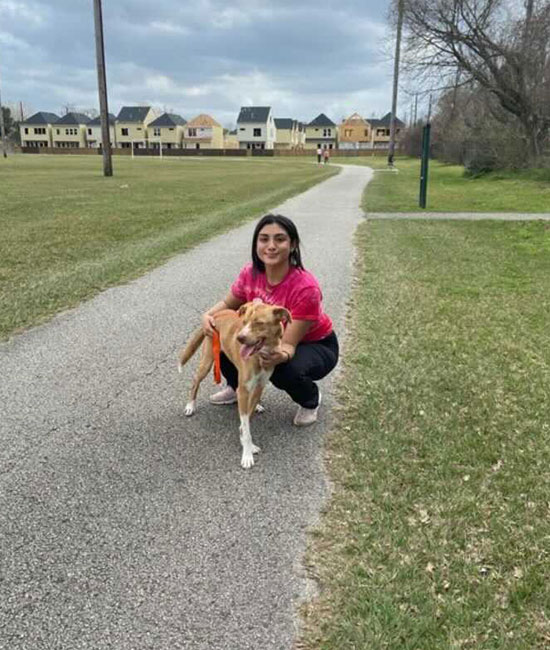 A woman in a pink shirt, possibly a veterinarian, crouches on a paved path, smiling and holding a brown dog on a leash. They are in a grassy park with houses and cloudy skies in the background.