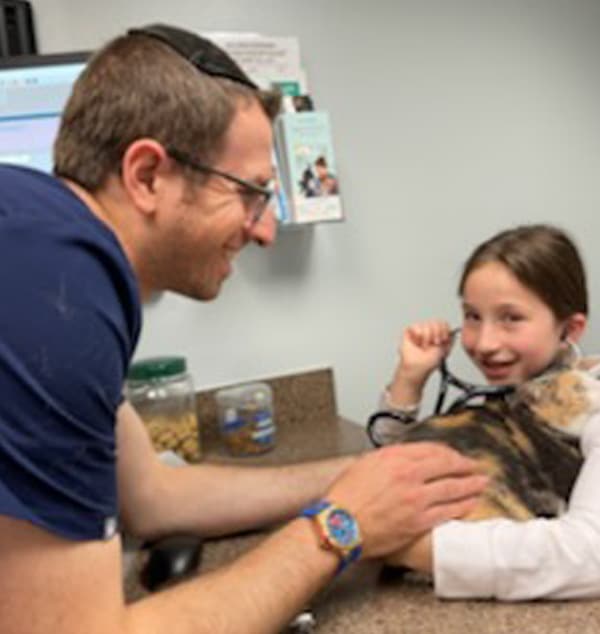 A man smiles while a young girl, aspiring to be a veterinarian, uses a stethoscope to listen to a cat on a vet’s exam table. The girl looks happy and the man appears encouraging.