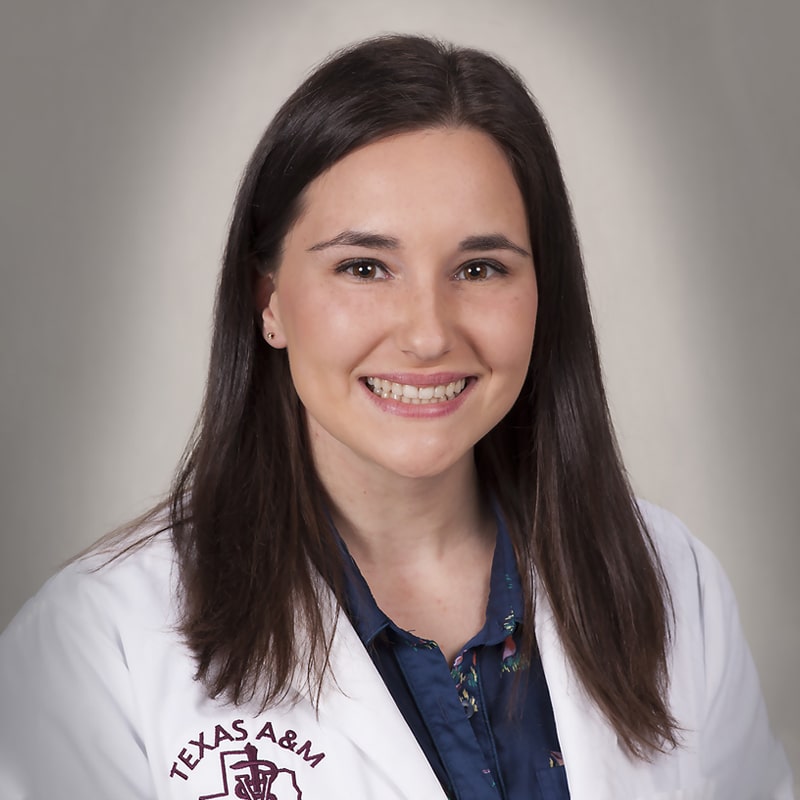 A woman with straight brown hair, wearing a white coat embroidered with "Texas A&M" and a dark blue blouse, smiles at the camera against a plain, light background, embodying the confidence of a dedicated veterinarian.