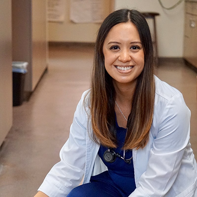 A woman with long brown hair, wearing a white lab coat and navy scrubs, sits smiling in a hallway as a veterinarian, with medical equipment and charts visible in the background.