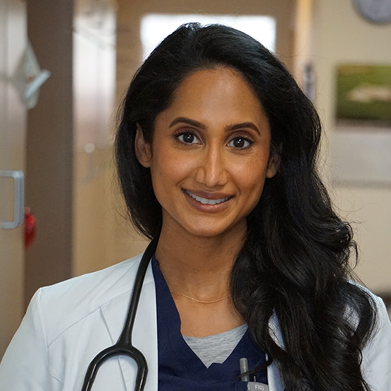 A woman with long dark hair, wearing a white coat and a stethoscope, smiles while standing in a hallway that appears to be in a veterinary clinic, showcasing her role as a dedicated veterinarian.