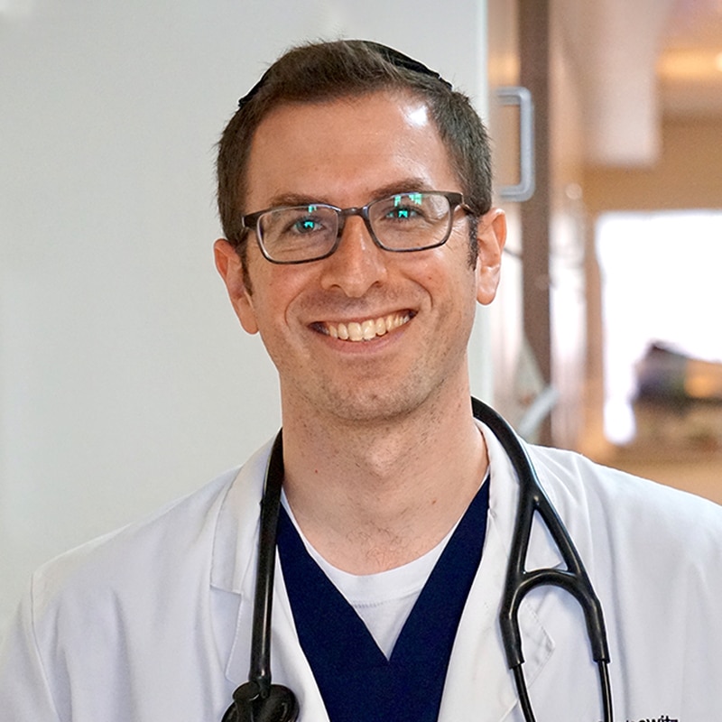 A smiling male vet wearing glasses, a white coat, and a stethoscope around his neck stands indoors in what appears to be a hospital or clinic hallway.