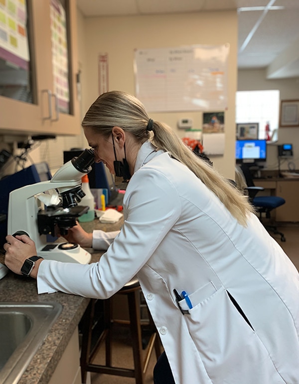 A veterinarian in a white lab coat examines a sample through a microscope in a laboratory setting. She has blonde hair tied back and wears a smartwatch and a mask around her neck, with computer screens and lab equipment in the background.