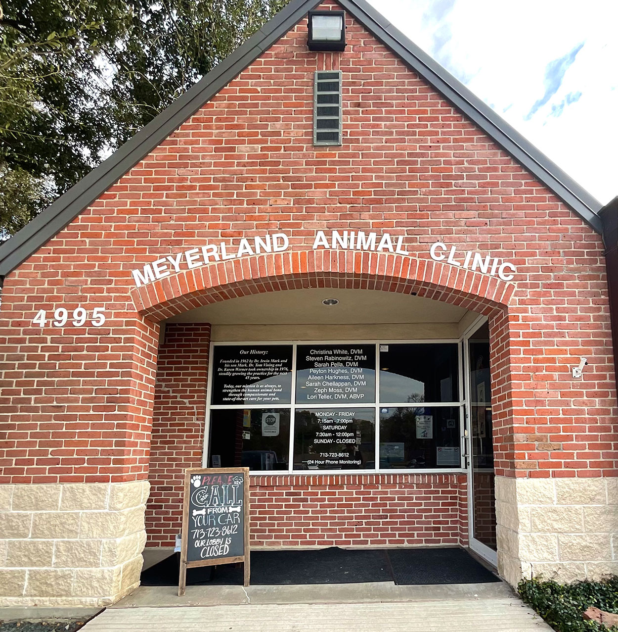 Brick building with "Meyerland Animal Clinic" written above the entrance. The number 4995 is on the left. A chalkboard sign stands by the door, and glass windows reflect trees and sky at this trusted veterinarian clinic.