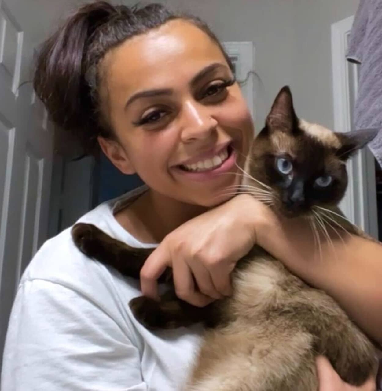 A woman with dark hair in a ponytail, possibly a veterinarian, smiles while holding a blue-eyed Siamese cat. The woman wears a white shirt, and they are indoors, standing by a doorway.