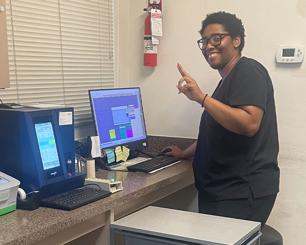 A person wearing black scrubs stands and smiles at the camera while using a desktop computer in an office with medical equipment and supplies. They are holding up one finger in a positive gesture.