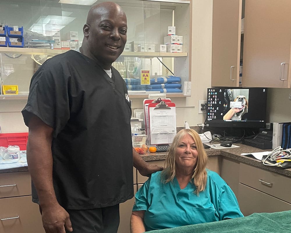 A smiling healthcare worker in black scrubs stands next to a seated woman in a teal medical gown in a clinic room, with medical equipment, supplies, and a computer visible in the background.