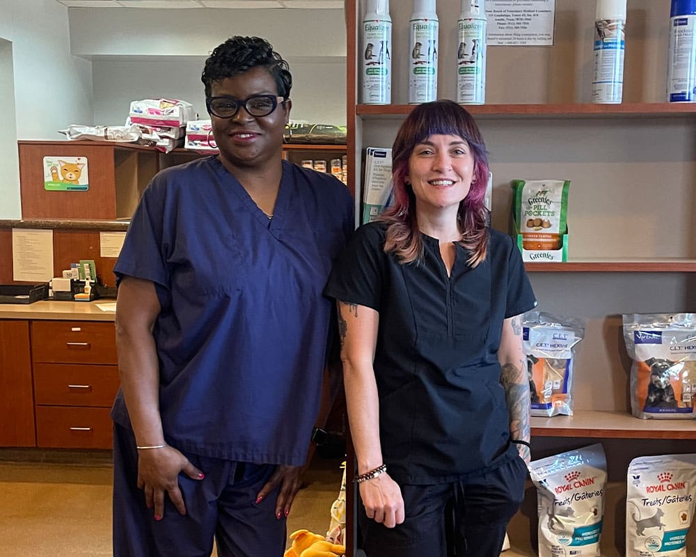 Two women wearing dark scrubs stand smiling in a veterinary clinic, with bags of pet food and supplies displayed on shelves behind them.