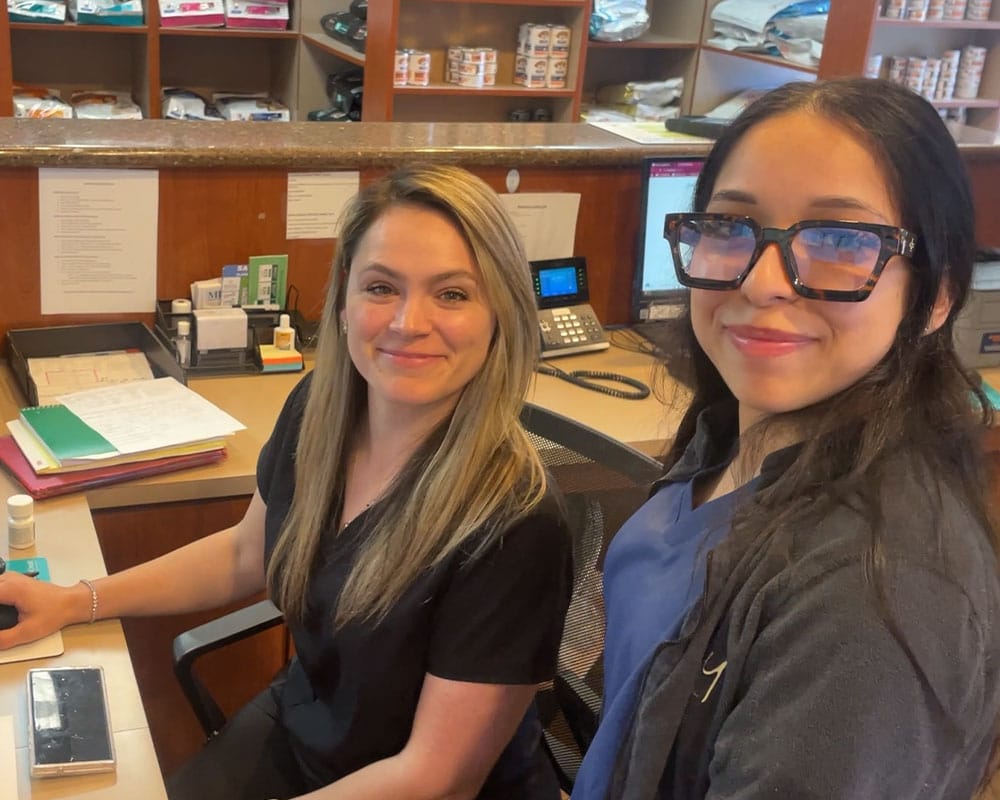 Two women wearing scrubs smile at the reception desk of a veterinary clinic. Shelves with pet products and paperwork are visible in the background, along with a computer and office supplies on the desk.