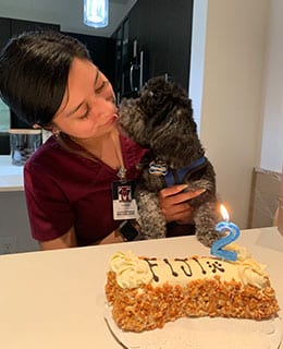 A veterinarian in scrubs holds a small black dog as they touch noses. In front of them is a cake with "Fiji" written on it and a blue number 2 candle, celebrating the dog's birthday.