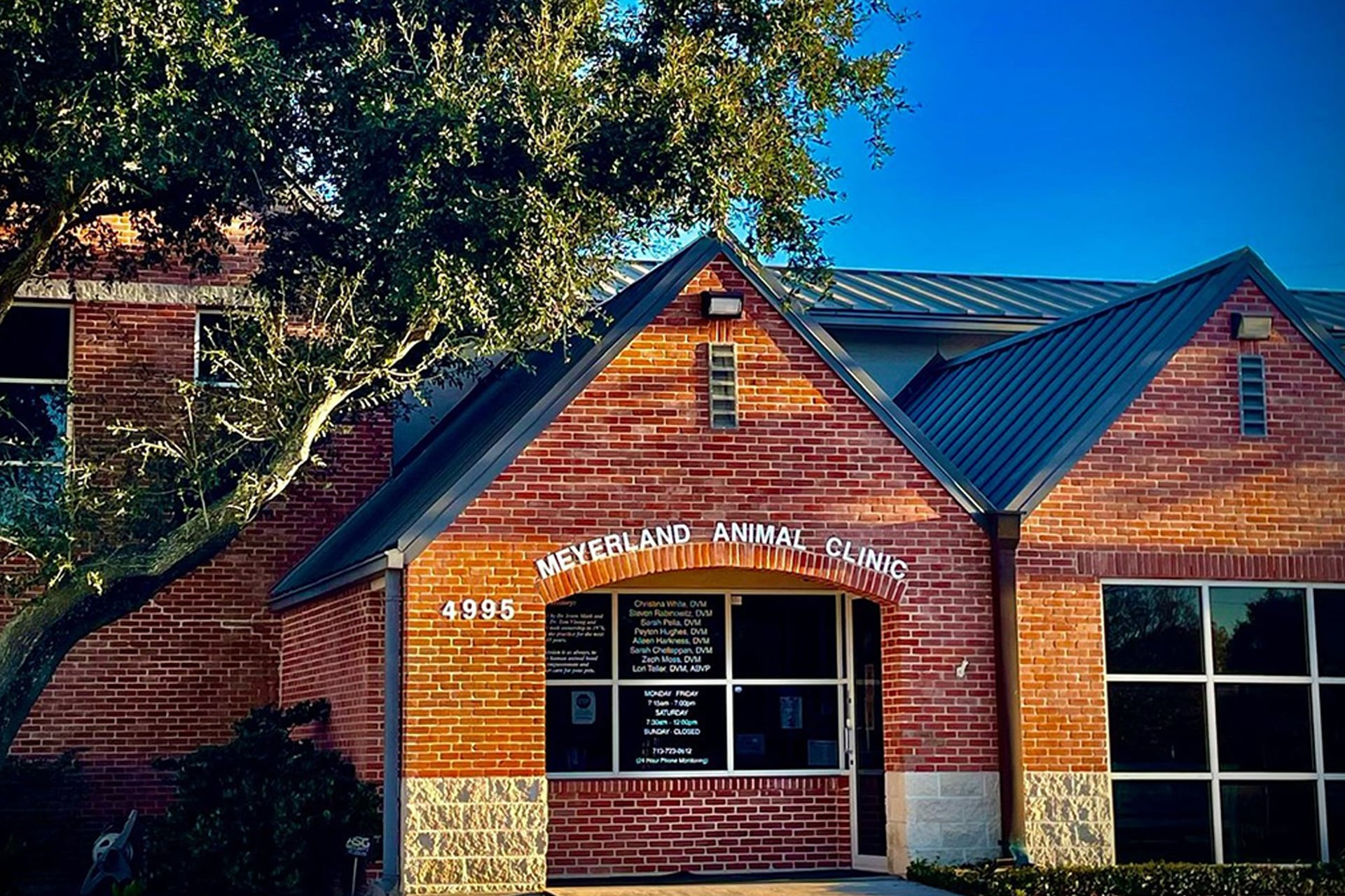 A red-brick building with a peaked metal roof houses the Meyerland Animal Clinic, where a caring veterinarian team welcomes pets. The entrance is shaded by a large tree, and the address number 4995 is displayed above the doorway.