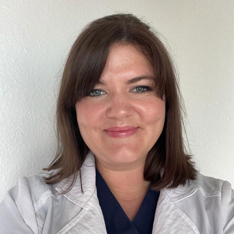A woman with shoulder-length brown hair and blue eyes smiles at the camera. She is wearing a white lab coat over a dark top, standing against a plain white background—a dedicated veterinarian ready to care for her patients.