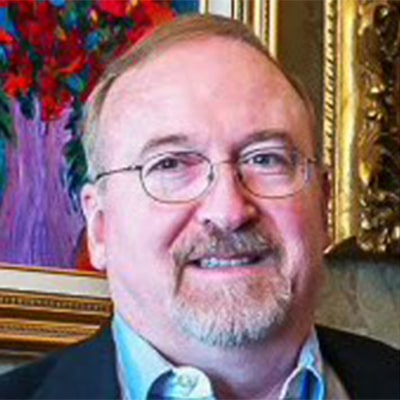 A middle-aged man with glasses, a short beard, and a mustache—dressed in a suit jacket—smiles at the camera. This veterinarian stands in front of a colorful painting and a gold-framed picture.