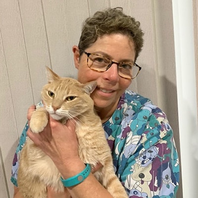 A smiling veterinarian wearing glasses and a colorful shirt with animal prints holds a light orange cat in their arms.